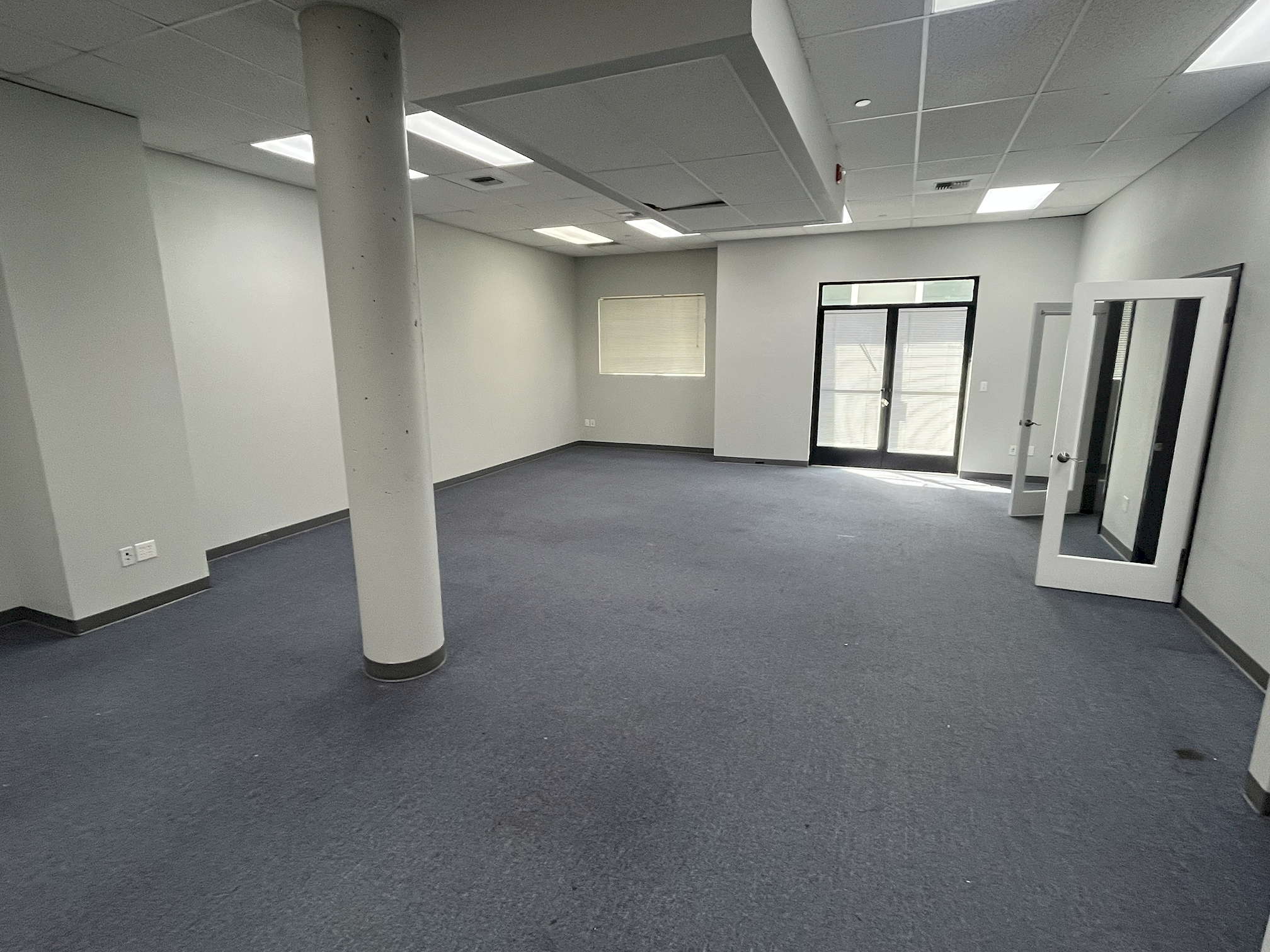 SDG pre-construction 1 A daytime photo of a plain office with white walls and ceiling, slate grey carpet, and an off-center pillar. At the back is a window and a pair of glass doors, and along the right wall are a couple plain doors with large windows in them.