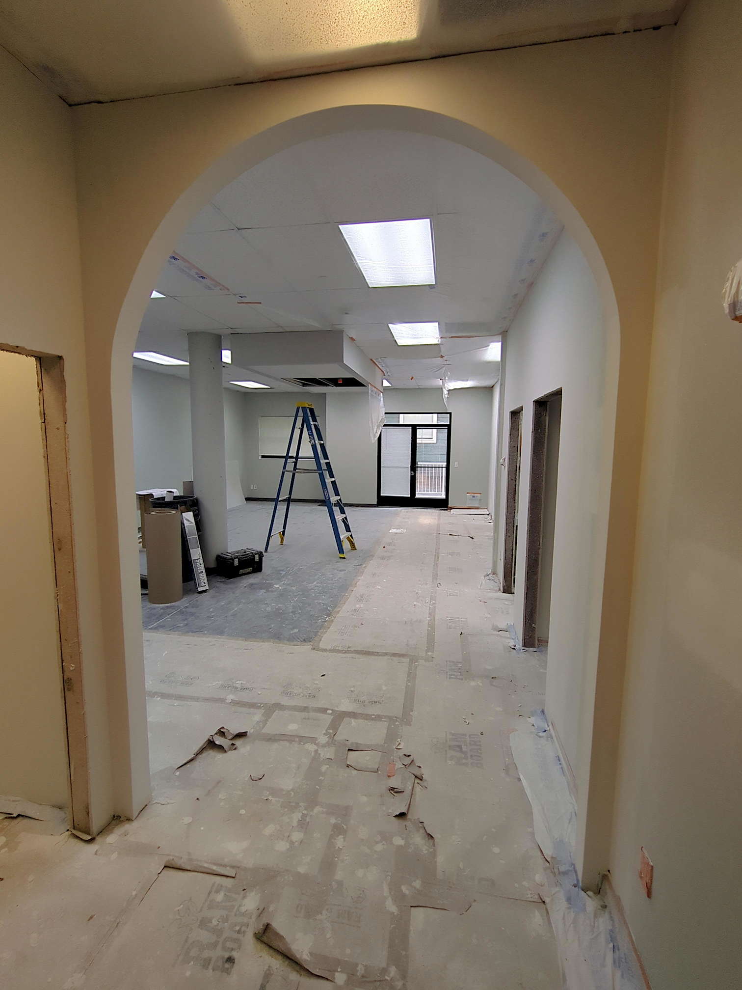 SDG pre-construction 9 A view through a tall rounded arch of a white-walled office space in the process of remodeling. The floor is covered in cardboard and platic tarps, along with some of the ceiling being covered in plastic. A ladder reaches up to a dropped area of the ceiling, and to the left are construction materials piled around a pillar.